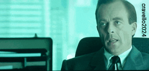a man in a suit and tie sits in front of a screen that says critello2024 on it