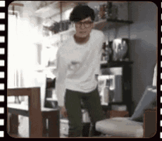 a man wearing glasses is standing in a living room in front of a chair .