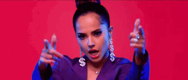 a woman wearing a purple jacket and earrings with dollar signs on them is making a peace sign with her hands .