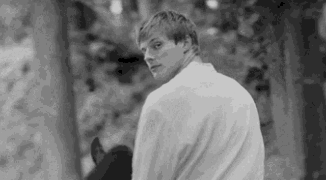a man in a white shirt is standing in the woods holding a hat .