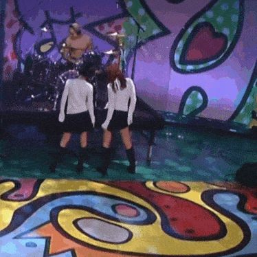 two women are standing on a stage with a man playing drums in the background