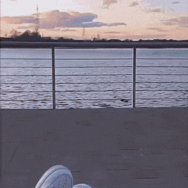 a person 's feet are visible in front of a railing overlooking a body of water at sunset
