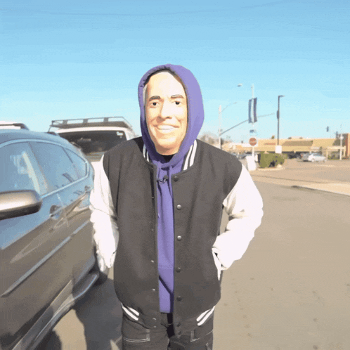 a man wearing a mask and a varsity jacket is standing in front of a car