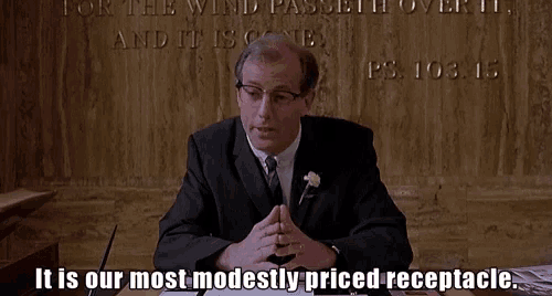 a man in a suit and tie is sitting at a desk with the words it is our most modestly priced receptacle