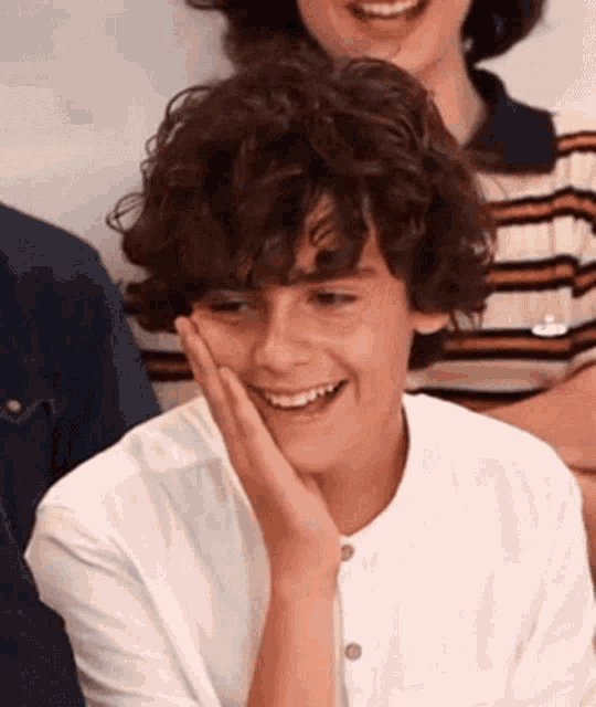 a young boy with curly hair is smiling with his hand on his chin .