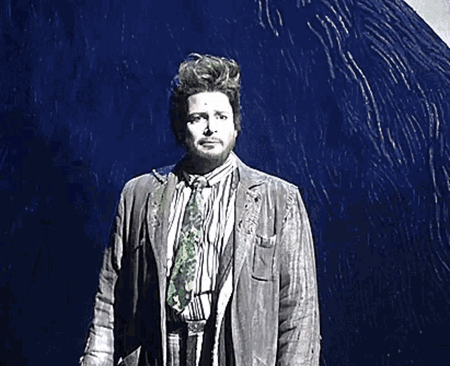 a man with a beard wearing a jacket and tie is standing in front of a blue wall .