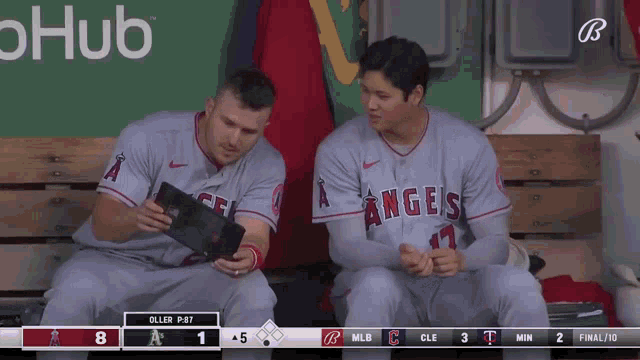 two angels baseball players sit on a bench looking at a tablet