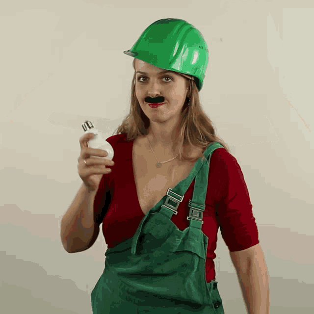 a woman wearing overalls and a green hard hat holds a light bulb