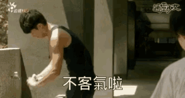 a man in a tank top is washing his hands in a sink while another man looks on .