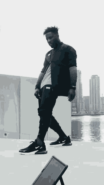 a man wearing a black jacket and black nike shoes stands in front of a body of water