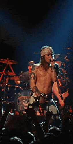 a shirtless man sings into a microphone in front of a drum set that says guns n ' roses