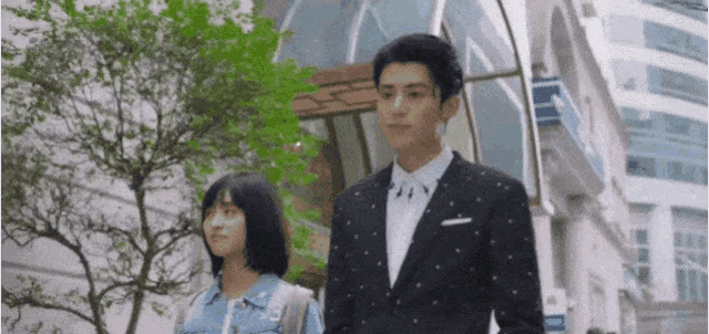 a man in a suit and a woman in a denim jacket are standing next to each other on a city street .