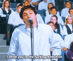 a man singing into a microphone with the words " i think you 're the fucking antichrist " above him