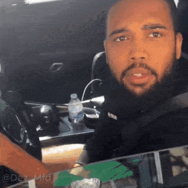 a man with a beard is sitting in a car with a bottle of water in the passenger seat