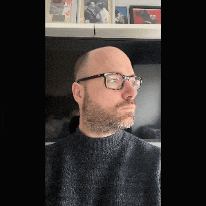 a bald man with glasses and a beard stands in front of a shelf with pictures on it