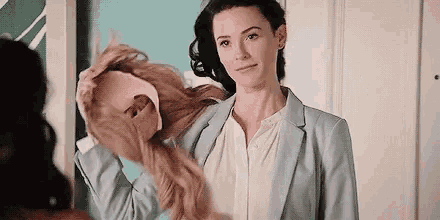 a woman in a suit is holding a wig in her hand while another woman looks on .