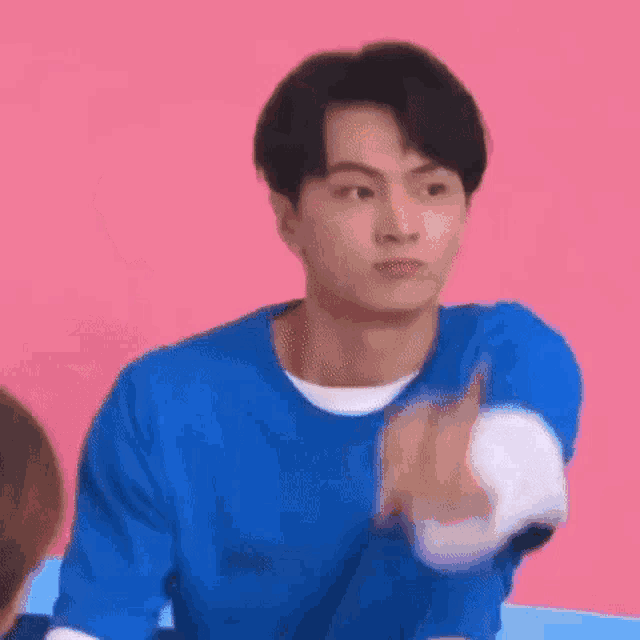 a young man in a blue shirt is making a funny face while sitting in front of a pink wall .