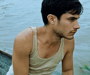 a man in a tan tank top sits on a boat in the water