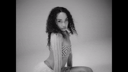 a black and white photo of a woman sitting on the floor wearing a bikini top and skirt .