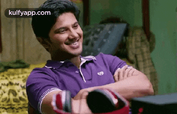 a man in a purple shirt is smiling with his arms crossed while sitting on a bed .