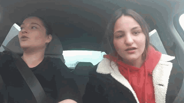 two women are sitting in a car one wearing a red hoodie