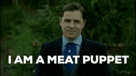 a man in a suit and tie is standing in front of a forest and says `` i am a meat puppet '' .