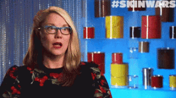 a woman wearing glasses stands in front of a wall of cans with the hashtag #skinwars on the bottom