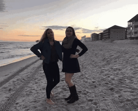 two women standing on a sandy beach with a sunset in the background