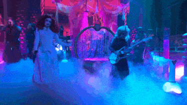 a woman in a white dress is standing in a foggy room with a man playing a guitar