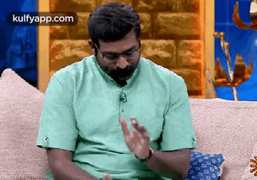 a man with a beard and glasses is sitting on a couch with his hands up .