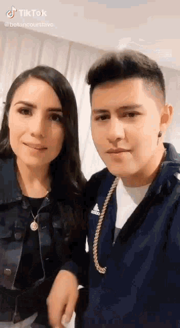 a man and a woman are posing for a picture together . the man is wearing a blue adidas jacket .