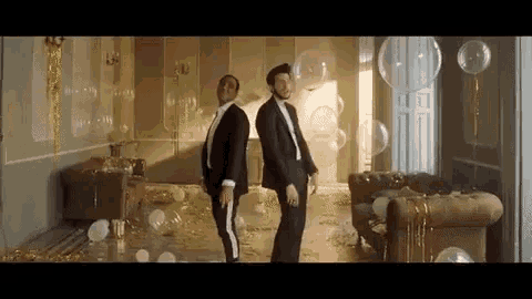 two men in suits are dancing in a room with balloons and confetti on the floor .