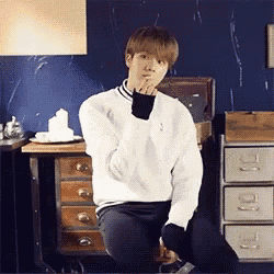 a young man in a white sweater is sitting on a stool with his hand on his chin .