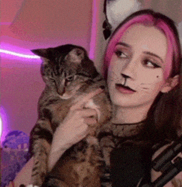 a woman with pink hair is holding a cat with a cat face paint on her face .