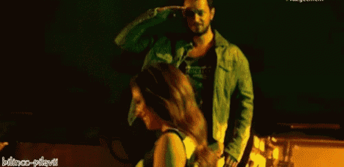 a man in a green jacket is standing next to a woman in a black tank top in a dark room .