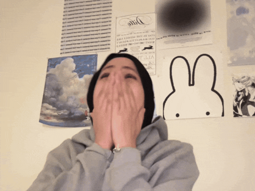 a person covering their mouth with their hands in front of a wall with posters and a drawing of a bunny