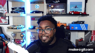a man wearing glasses and a black shirt is sitting in front of a shelf full of toy cars