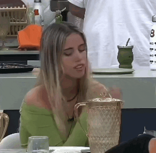 a woman in a green top sits at a table with a pitcher and a glass
