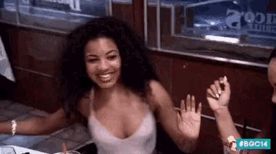 a woman in a white tank top is sitting at a table waving her hand .