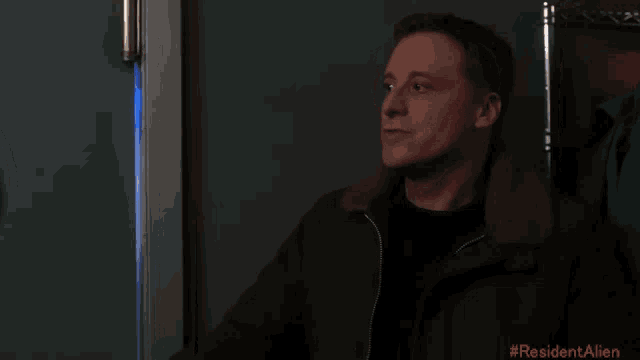 a man in a brown jacket is standing in a dark room with #resident alien written on the bottom