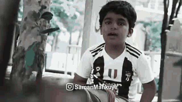 a young boy wearing a juventus jersey is sitting next to a tree and talking to someone .