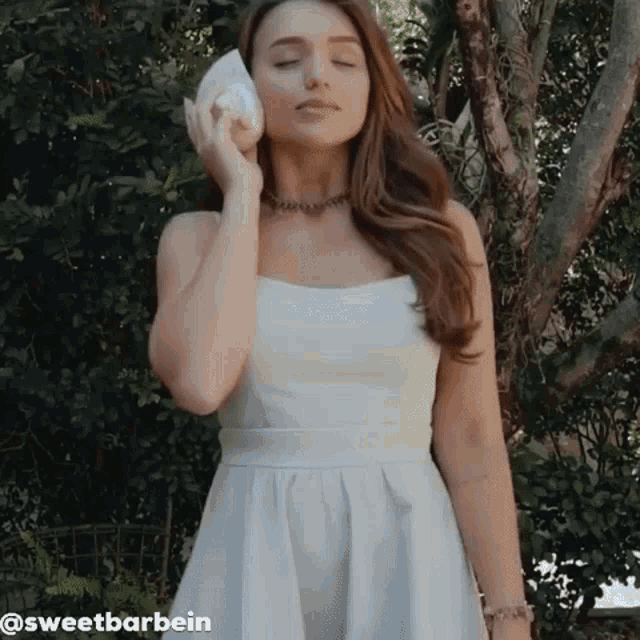 a woman in a white dress is holding a seashell to her ear .