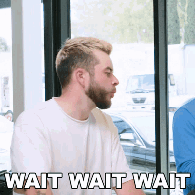 a man with a beard is sitting in front of a window with wait wait wait written below him