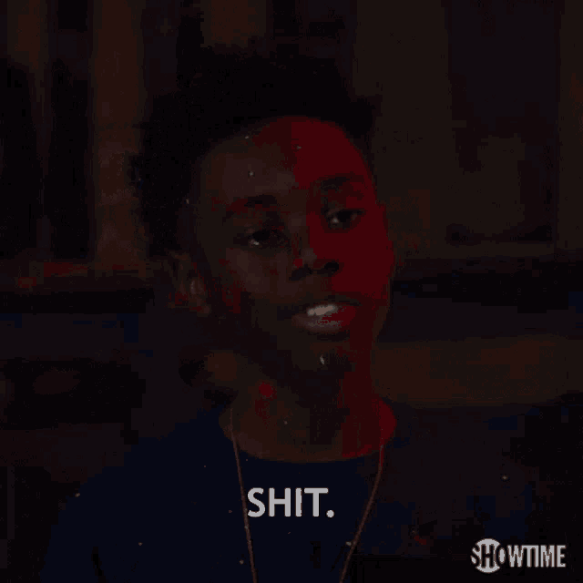 a young man in a blue shirt is standing in a dark room and says shit .