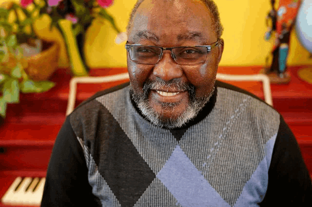 a man wearing glasses and a plaid sweater smiles in front of a piano