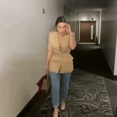 a woman in a yellow jacket and jeans is walking down a hallway holding a bag .