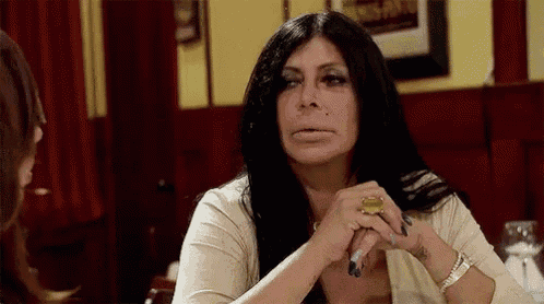 a woman with long black hair is sitting at a table with her hands folded and smoking a cigarette .