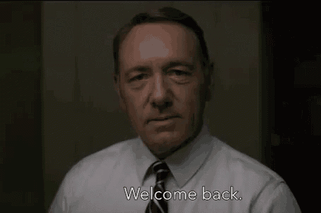 a man in a white shirt and tie is standing in front of a wall and saying `` welcome back '' .