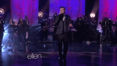 a man in a suit is singing into a microphone on a stage in front of a sign that says ellen