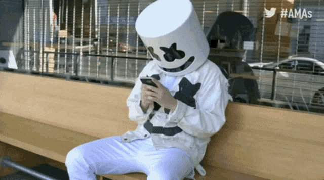 a person wearing a marshmallow mask sitting on a bench looking at their phone